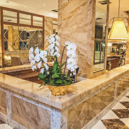 Windsor Marapendi Hotel Rio de Janeiro Ngoại thất bức ảnh The photo depicts a luxurious hotel lobby featuring a polished marble reception desk. There are elegant white orchids arranged in a gold pot on the counter, adding a touch of sophistication. The surrounding interior showcases marble walls, stylish li