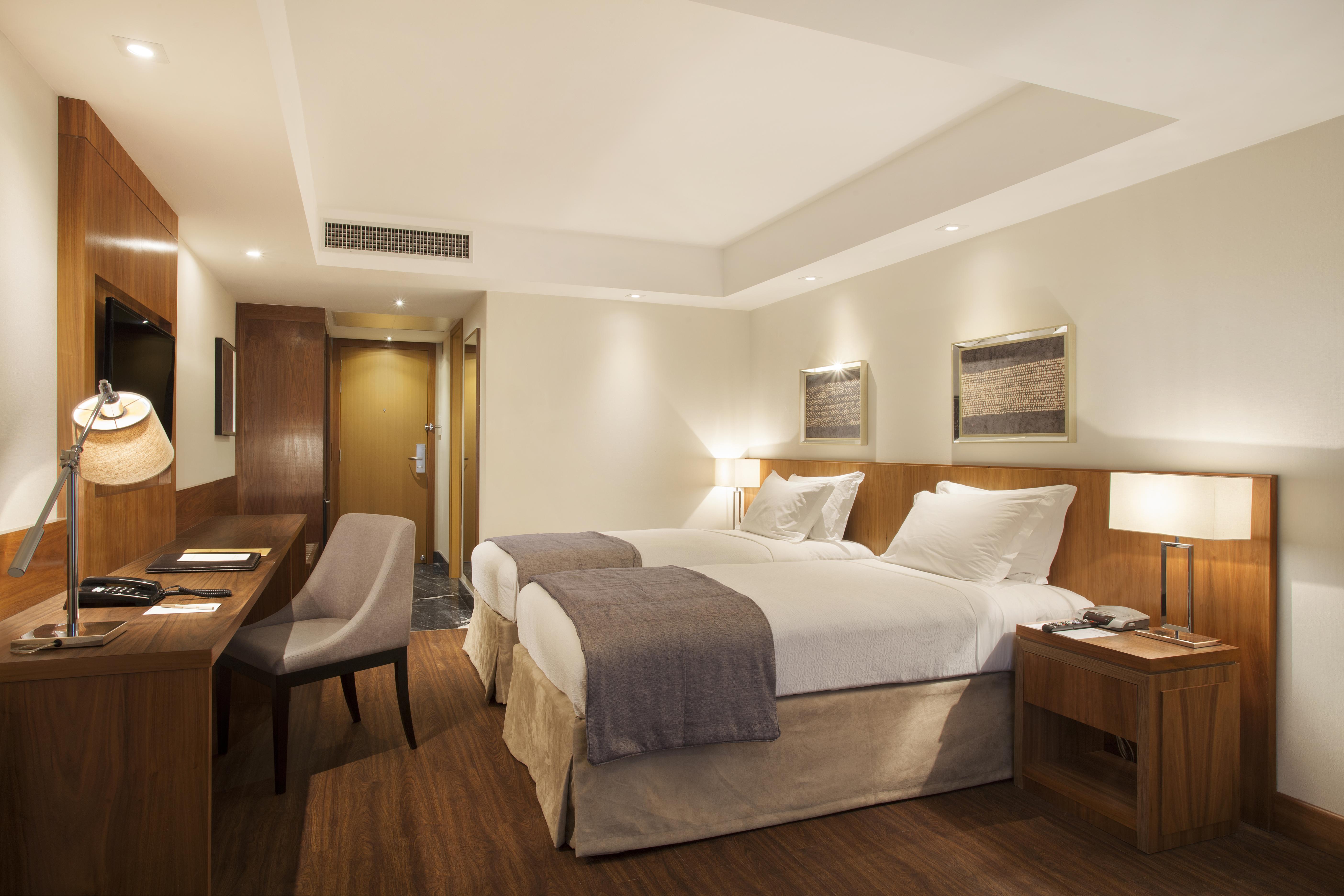 Windsor Marapendi Hotel Rio de Janeiro Ngoại thất bức ảnh The photo displays a modern hotel room featuring two twin beds with crisp white linens and gray throw blankets. The room has a clean and minimalist design, with wooden accents, including a bedside table and a desk. To the left, there's a small desk a