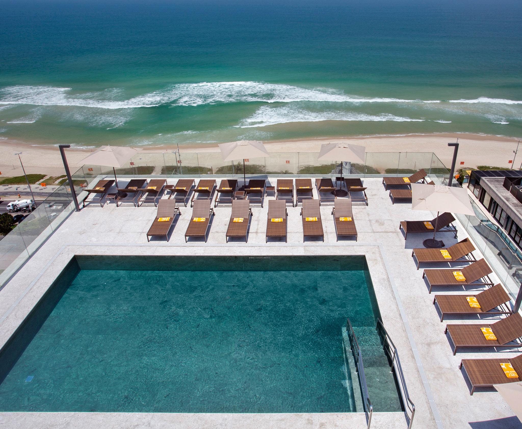 Windsor Marapendi Hotel Rio de Janeiro Ngoại thất bức ảnh The photo shows a rooftop area with a swimming pool and several lounge chairs arranged around it. The pool has a clear blue surface, and the lounge chairs are positioned in a neat row, some with yellow cushions. In the background, you can see a beaut