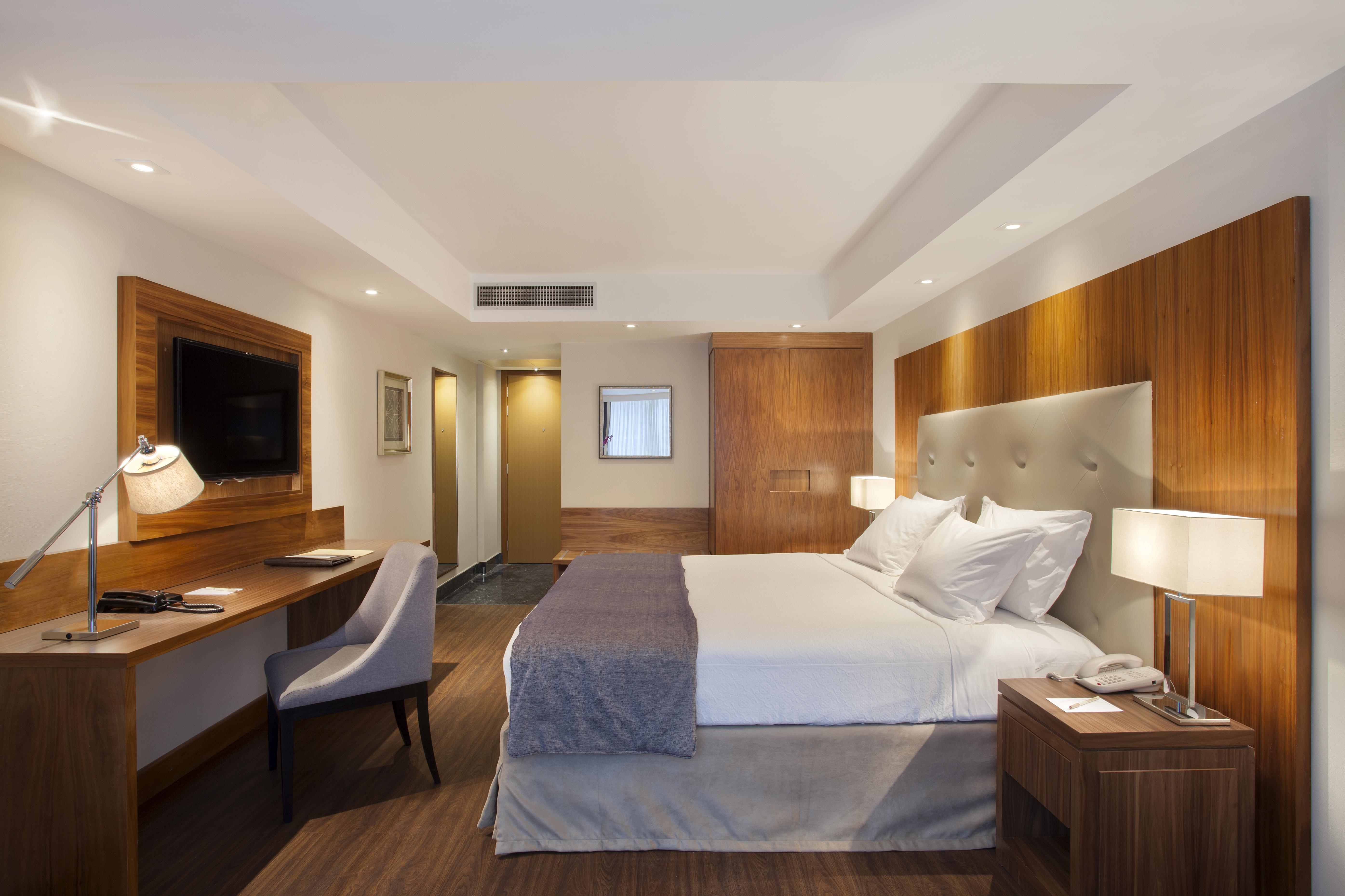 Windsor Marapendi Hotel Rio de Janeiro Ngoại thất bức ảnh The photo shows a modern hotel room featuring a large bed with white and gray bedding. The headboard is upholstered in light fabric. There are two bedside lamps on either side of the bed, providing warm lighting. A television is mounted on the wall o
